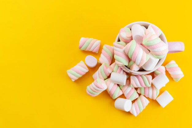 Eine Draufsicht färbte köstliche Marshmallows innerhalb der weißen Tasse auf gelbem Schreibtisch, süße Zuckerfarbe