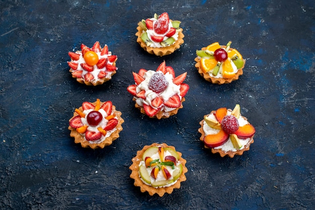 Eine Draufsicht verschiedene kleine Kuchen mit Sahne und frisch geschnittenen Früchten auf dem blauen Backgound-Obstkuchen-Keks