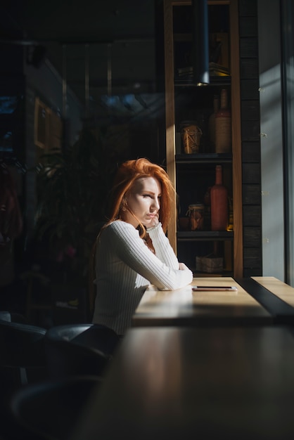 Eine einsame junge Frau, die nahe dem Fenster mit Handy auf Tabelle sitzt