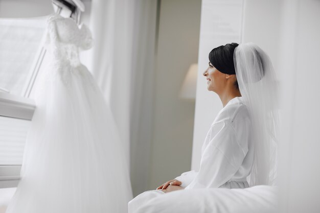 Eine elegante und schöne Braut zu Hause, die in einem Bett nahe Fenster sitzt