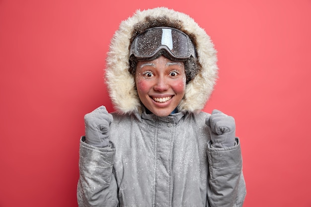 Eine emotional positive Frau hebt die Fäuste und kann es kaum erwarten, bis das Snowboarden fröhliche Gefühle zum Ausdruck bringt. Genießt die Winterzeit und die aktive Erholung trägt eine warme Jacke und eine Skibrille. Fröhliche Skifahrerin