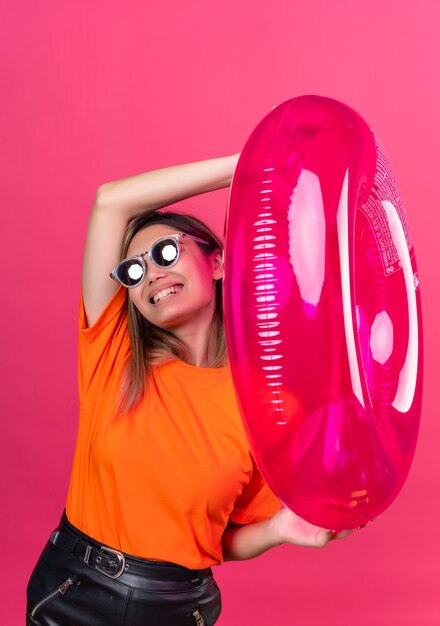 Eine erfreute junge Frau in einem orangefarbenen T-Shirt, das eine Sonnenbrille trägt, während sie rosa aufblasbaren Ring auf einer rosa Wand hält