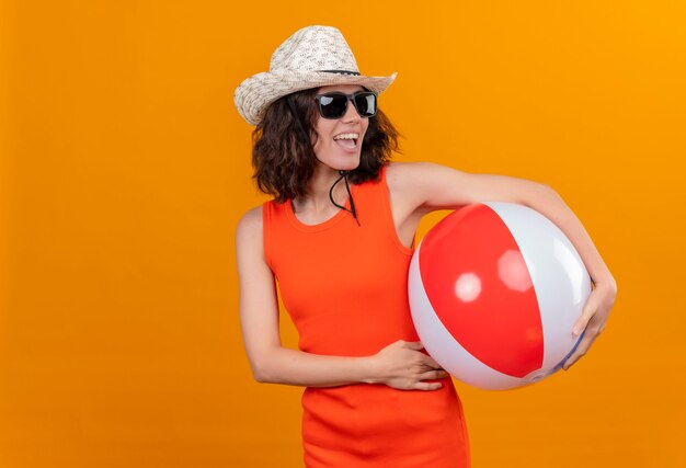 Eine erfreute junge Frau mit kurzen Haaren in einem orangefarbenen Hemd, das Sonnenhut und Sonnenbrille hält, die aufblasbare Kugel schauende Seite hält