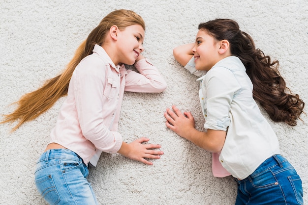 Eine erhöhte Ansicht von zwei lächelnden Mädchen, die auf dem Teppich liegen, der einander betrachtet