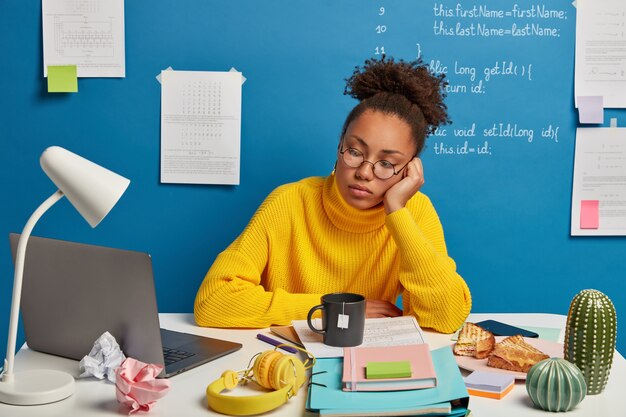 Eine ernsthafte konzentrierte Studentin nutzt den Online-Bildungsdienst, sieht sich ein Schulungswebinar oder einen Kurs auf einem Laptop an, hat viele Dinge auf dem Tisch und trinkt Tee