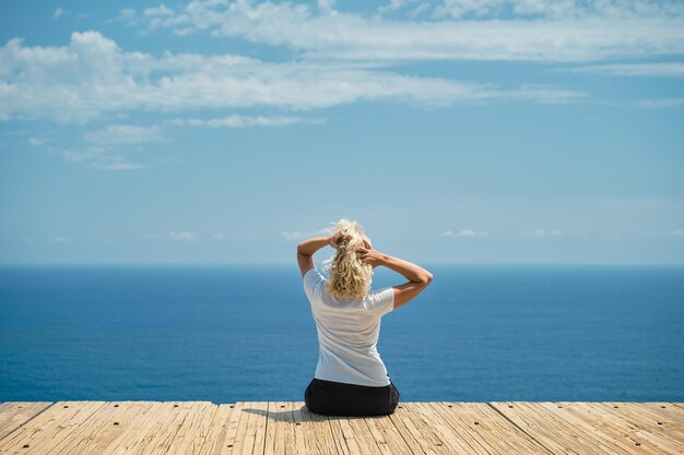 Eine erwachsene schöne Frau freut sich über einen warmen Tag. Die Blondine sitzt vor dem Hintergrund einer herrlichen Meereslandschaft auf einer Reise durch die griechischen Inseln, einem berühmten Ort für Urlaub und Reisen