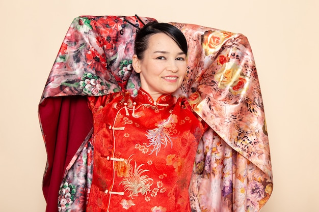 Eine exquisite japanische Geisha der Vorderansicht im traditionellen roten japanischen Kleid, das mit dem eleganten Lächeln des Blumendesigngewebes auf der cremefarbenen Hintergrundzeremonie Japan aufwirft