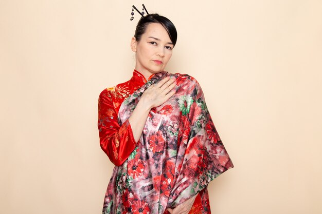 Eine exquisite japanische Geisha der Vorderansicht im traditionellen roten japanischen Kleid, das mit dem von der Blume entworfenen Gewebe elegant auf der Cremehintergrundzeremonie Japan aufwirft