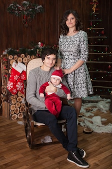 Eine familie mit einem kleinen kind im weihnachtsmannkostüm zu hause vor einem kamin auf einem schaukelstuhl