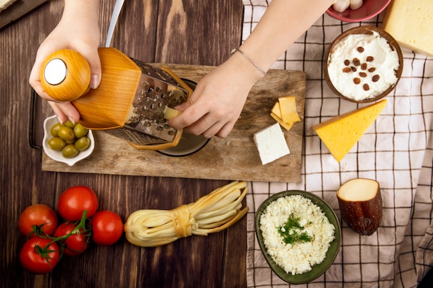Eine Frau, die Käse auf einem Holzbrett mit frischen Tomaten der eingelegten Oliven und verschiedenen Käsesorten auf Holz Draufsicht reibt
