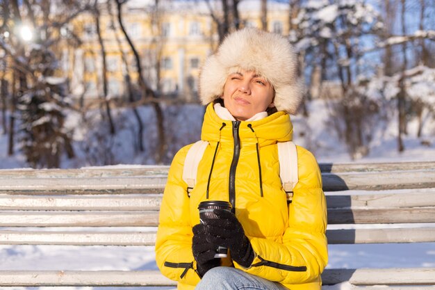 Eine Frau im Winter in warmer Kleidung in einem schneebedeckten Park an einem sonnigen Tag sitzt auf einer Bank und friert vor Kälte, ist im Winter unglücklich, hält Kaffee allein