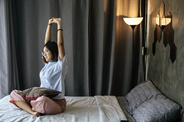 Eine Frau in einem weißen Hemd sitzt auf dem Bett und hebt beide Arme.