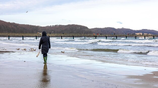 Eine Frau in Winterjacke und Stiefeln geht am Meer entlang