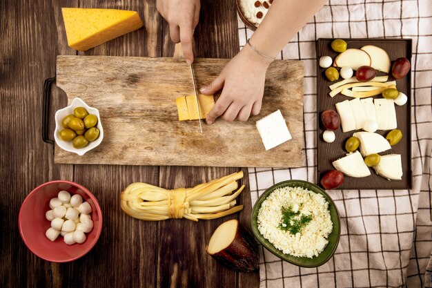 Eine Frau schneidet holländischen Käse auf einem Holzschneidebrett mit eingelegten Oliven und verschiedenen Käsesorten auf rustikaler Draufsicht