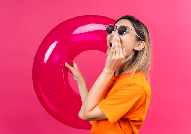 Eine freudige hübsche junge Frau in einem orangefarbenen T-Shirt mit Sonnenbrille, die jemanden anruft, während sie rosa aufblasbaren Ring an einer rosa Wand hält