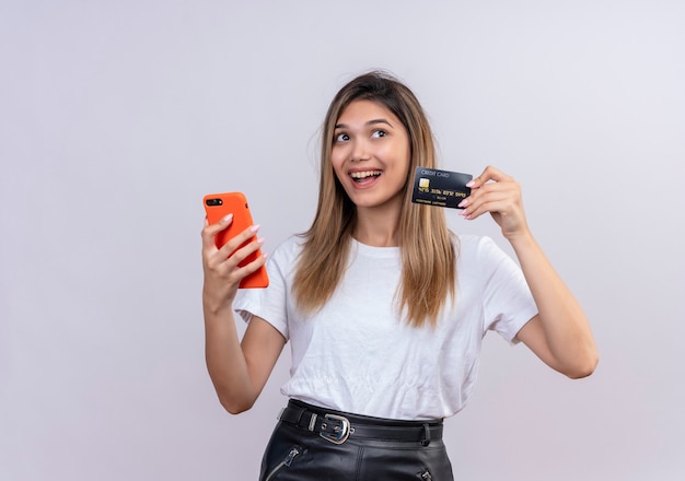 Eine fröhliche junge Frau im weißen T-Shirt, das Kreditkarte zeigt, während Handy auf einer weißen Wand hält