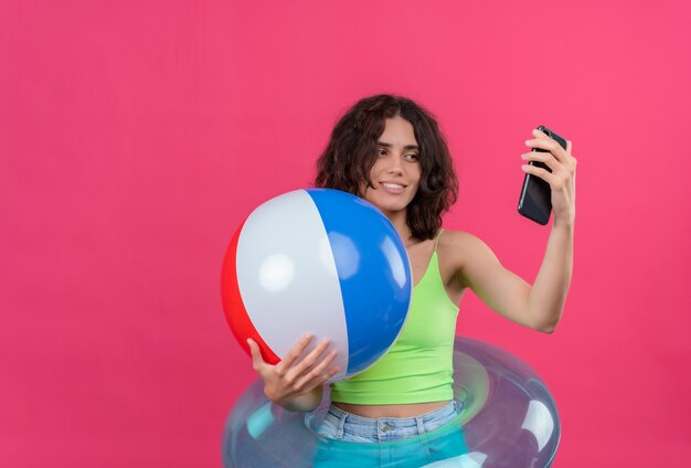 Eine fröhliche junge Frau mit kurzen Haaren im grünen Erntedach, die lächelnd und aufblasbaren Ball hält, der selfie mit Handy nimmt