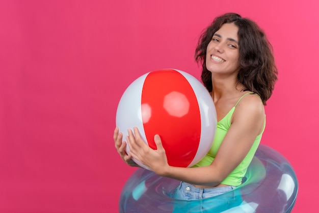 Eine fröhliche junge Frau mit kurzen Haaren in der grünen Erntespitze, die lächelnd und aufblasbaren Ball hält