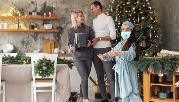 Eine glückliche Familie trägt wegen des COVID-19-Coronavirus in der Nähe des Weihnachtsbaums medizinische Masken. Weihnachtsferien.