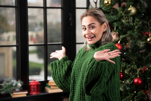 Kostenloses Foto eine glückliche frau, die einen grünen pullover trägt, der nahe dem weihnachtsbaum aufwirft.