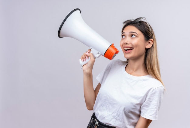 Eine glückliche hübsche junge Frau im weißen T-Shirt, das Sonnenbrille auf ihrem Kopf trägt und durch Megaphon auf einer weißen Wand spricht