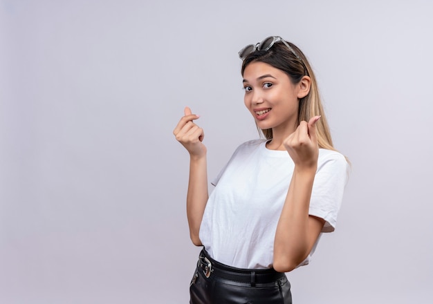 Eine glückliche hübsche junge Frau im weißen T-Shirt, das Sonnenbrille auf ihrem Kopf trägt, zeigt Geste für Geld auf einer weißen Wand