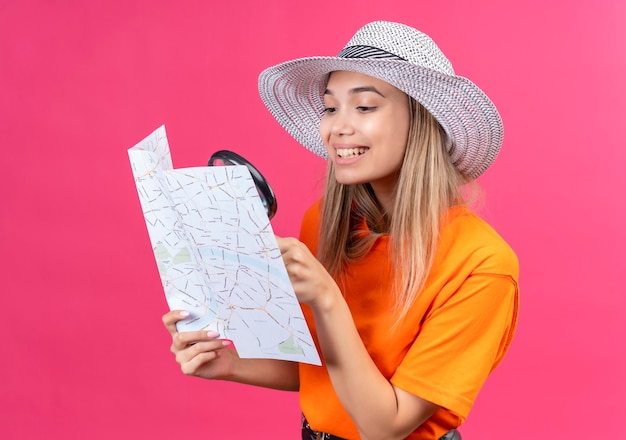 Eine glückliche hübsche junge Frau in einem orange T-Shirt, das Sonnenhut trägt, der eine Karte mit Lupe auf einer rosa Wand betrachtet