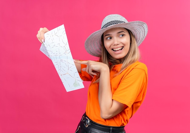 Eine glückliche hübsche junge Frau in einem orangefarbenen T-Shirt, das einen lächelnden Sonnenhut trägt, während er auf eine Karte zeigt und Seite auf einer rosa Wand schaut