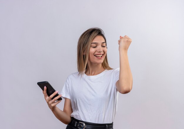 Eine glückliche reizende junge Frau im weißen T-Shirt, das geballte Faust hebt, während Handy auf einer weißen Wand hält