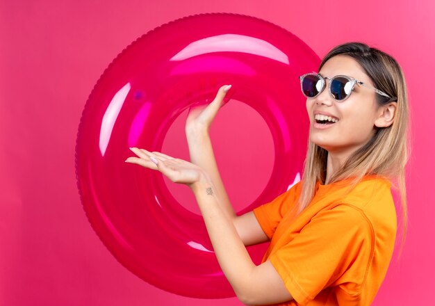 Eine glückliche reizende junge Frau in einem orangefarbenen T-Shirt, das eine Sonnenbrille trägt, die lächelt und schaut, während sie rosa aufblasbaren Ring auf einer rosa Wand hält