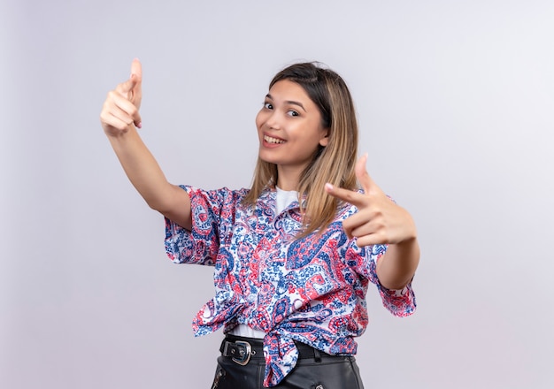 Eine glückliche schöne junge Frau, die ein mit Paisley bedrucktes Hemd trägt, das mit Zeigefinger zeigt, während sie es auf einer weißen Wand betrachtet