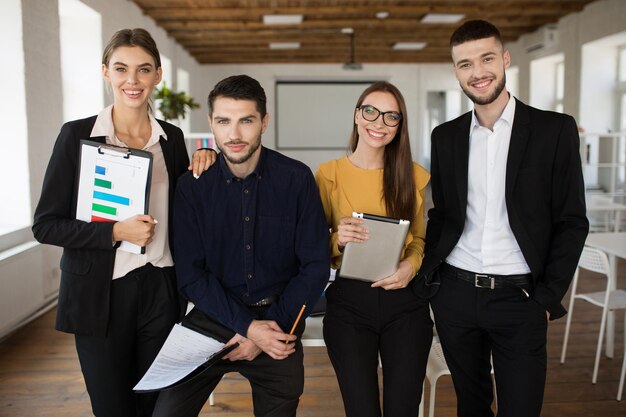 Eine Gruppe junger Geschäftsleute in klassischen Anzügen, die glücklich zusammen in die Kamera schauen, während sie Zeit bei der Arbeit in einem modernen Büro verbringen