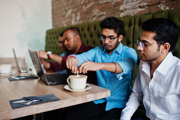 Eine Gruppe von vier südasiatischen Männern posierte bei einem Geschäftstreffen im Café. Inder arbeiten mit Laptops zusammen und verwenden verschiedene Geräte, unterhalten sich und trinken Kaffee