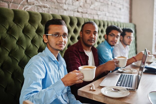 Eine Gruppe von vier südasiatischen Männern posierte bei einem Geschäftstreffen im Café. Inder arbeiten mit Laptops zusammen und verwenden verschiedene Geräte, unterhalten sich und trinken Kaffee