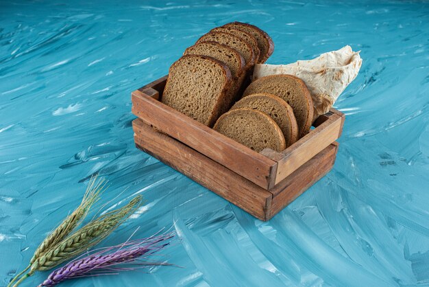 Eine Holzkiste voll geschnittenem frischem Schwarzbrot mit Weizenähren auf blauer Oberfläche