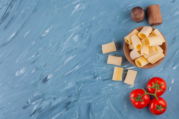 Eine Holzschale mit trockenen rohen Röhrennudeln mit frischen roten Tomaten und Gewürzen auf einem blauen Tisch.