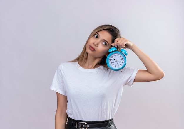 Eine hübsche junge Frau im weißen T-Shirt, die Uhr tickt Ton hört, während blauer Wecker auf einer weißen Wand hält