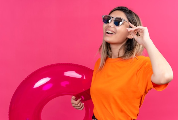 Eine hübsche junge Frau in einem orangefarbenen T-Shirt, das eine Sonnenbrille trägt, die lächelt und Hand auf Sonnenbrille hält, während rosa aufblasbaren Ring an einer rosa Wand hält