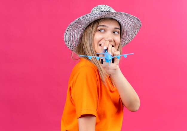 Eine hübsche junge Frau in einem orangefarbenen T-Shirt, das einen Sonnenhut trägt, der lächelt und vorwärts schaut, während er blaues Spielzeugflugzeug auf einer rosa Wand hält