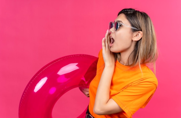 Kostenloses Foto eine hübsche junge frau in einem orangefarbenen t-shirt mit sonnenbrille, die jemanden anruft, während sie rosa aufblasbaren ring an einer rosa wand hält