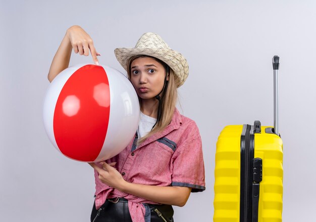 Eine hübsche reizende junge Frau, die rotes Hemd und Sonnenhut hält aufblasbaren Ball auf einer weißen Wand