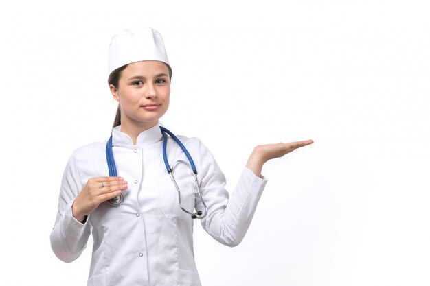 Eine junge Ärztin der Vorderansicht im weißen medizinischen Anzug und in der weißen Kappe mit blauem Stethoskop