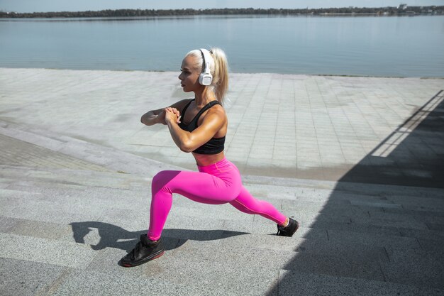 Eine junge athletische Frau im Hemd und in den weißen Kopfhörern, die trainieren, die Musik auf der Straße draußen zu hören. Ausfallschritte an einem sonnigen Tag. Konzept des gesunden Lebensstils, des Sports, der Aktivität, des Gewichtsverlusts.