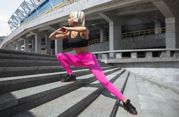 Eine junge athletische Frau im Hemd und in den weißen Kopfhörern, die trainieren, die Musik auf der Straße draußen zu hören. Ausfallschritte die Treppe hinauf. Konzept des gesunden Lebensstils, des Sports, der Aktivität, des Gewichtsverlusts.