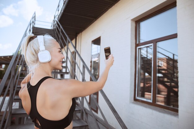 Eine junge athletische Frau im Hemd und in den weißen Kopfhörern, die trainieren, die Musik auf einer Treppe im Freien zu hören. Ein Selfie machen. Konzept des gesunden Lebensstils, des Sports, der Aktivität, des Gewichtsverlusts.
