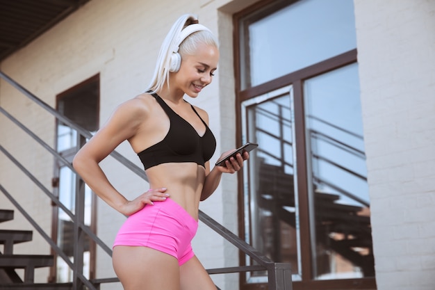 Eine junge athletische Frau im Hemd und in den weißen Kopfhörern, die trainieren, die Musik auf einer Treppe im Freien zu hören. Track auswählen oder chatten. Konzept des gesunden Lebensstils, des Sports, der Aktivität, des Gewichtsverlusts.