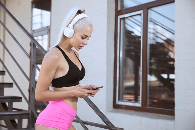 Eine junge athletische Frau im Hemd und in den weißen Kopfhörern, die trainieren, die Musik auf einer Treppe im Freien zu hören