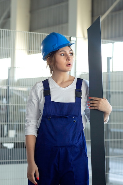 Eine junge attraktive Dame der Vorderansicht im blauen Bauanzug und im Helm, die das schwere metallische Detail während des Architekturbaus der Tagesgebäude halten