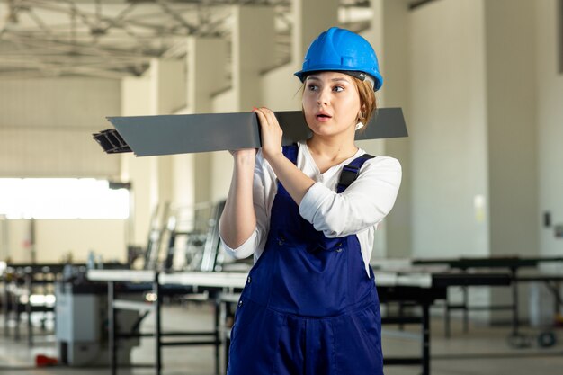 Eine junge attraktive Dame der Vorderansicht im blauen Bauanzug und im Helm, die schweres metallisches Detail während der tagsüber überraschten Gebäudearchitekturkonstruktion halten