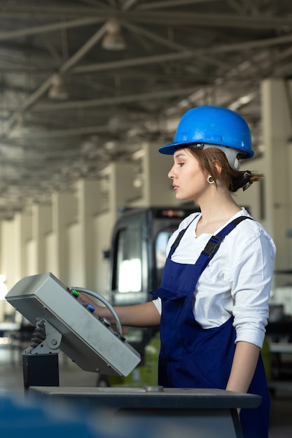 Eine junge attraktive Dame der Vorderansicht im blauen Bauanzug und in den Helm, die Maschinen im Hangar steuern, die während der Architektur des Tagesgebäudes arbeiten